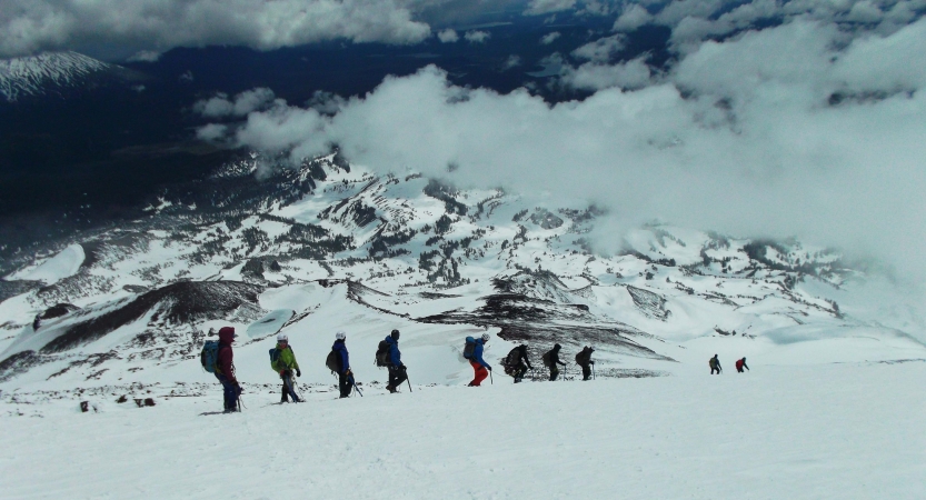 A line of people wearing snow gear, carrying backpacks and using trekking poles, hike through the snow of a mountainous landscape.